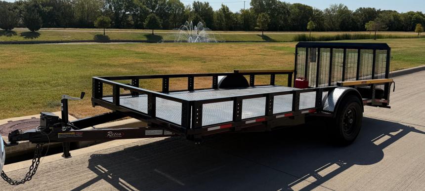 Fourteen-foot black single-axle Retco utility trailer with low diamond-plate sides, an open top, and a mesh ramp gate folded upright at the rear, parked on a concrete drive