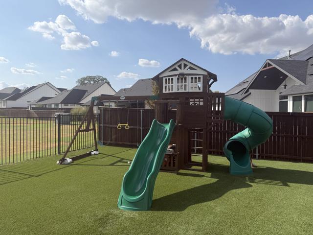 Large wooden backyard playset with a green tube slide, wave slide, and swing set standing on artificial turf before removal