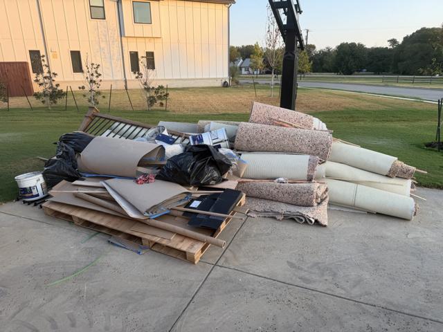 Driveway piled with remodeling debris including rolled carpet and padding, drywall sheets, lumber, cabinet doors, paint buckets, and black trash bags