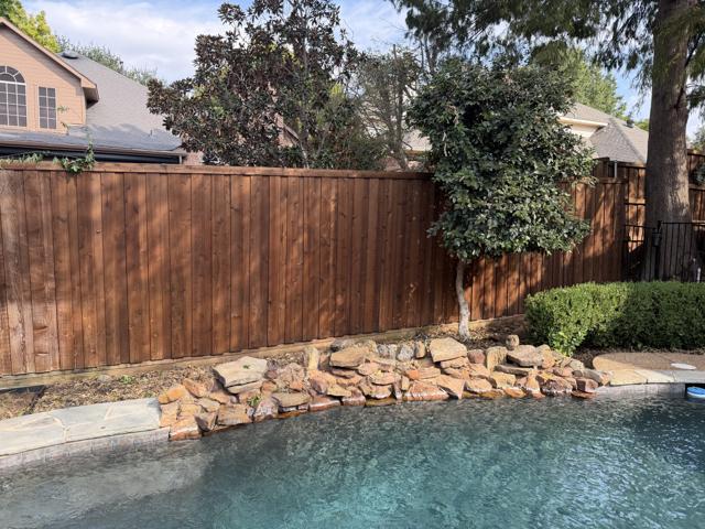 The same poolside bed after removal, with the trellis and vines gone, the stained privacy fence showing, and the rock border and trees left in place