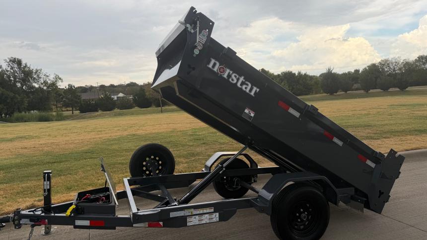 Black Norstar single-axle hydraulic dump trailer on a concrete driveway with its tall steel bed tipped up on the hydraulic cylinder, spare tire and battery box mounted on the tongue