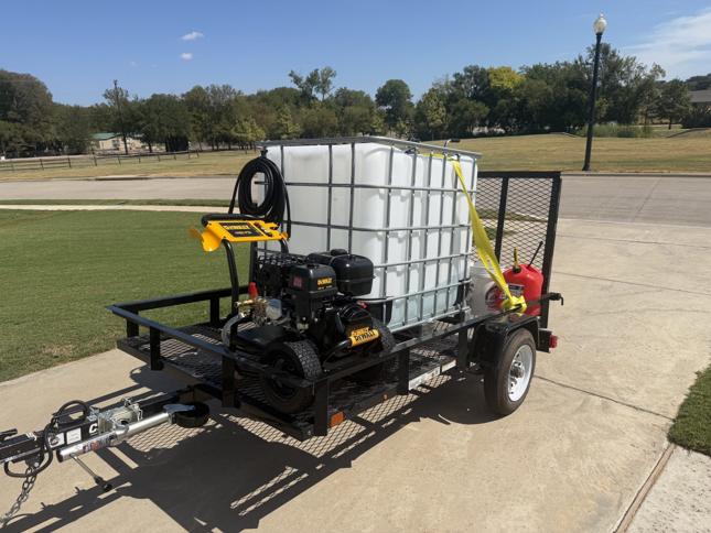Black single-axle utility trailer with a mesh floor and a ramp gate, carrying a white 275-gallon water tank, a yellow DeWalt gas pressure washer with its hose coiled on the handle, a fuel can, and a bucket strapped down, parked on a concrete driveway