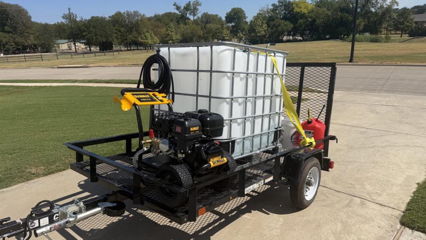 Black single-axle utility trailer with a mesh floor and a ramp gate, carrying a white 275-gallon water tank, a yellow DeWalt gas pressure washer with its hose coiled on the handle, a fuel can, and a bucket strapped down, parked on a concrete driveway